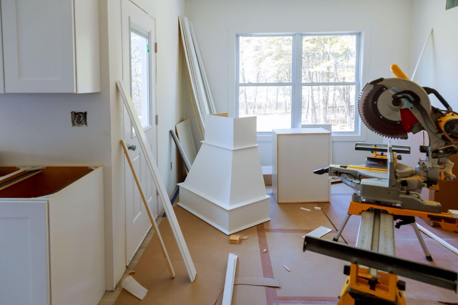 A kitchen under construction with new cabinets, a range hood, and a miter saw on the floor.