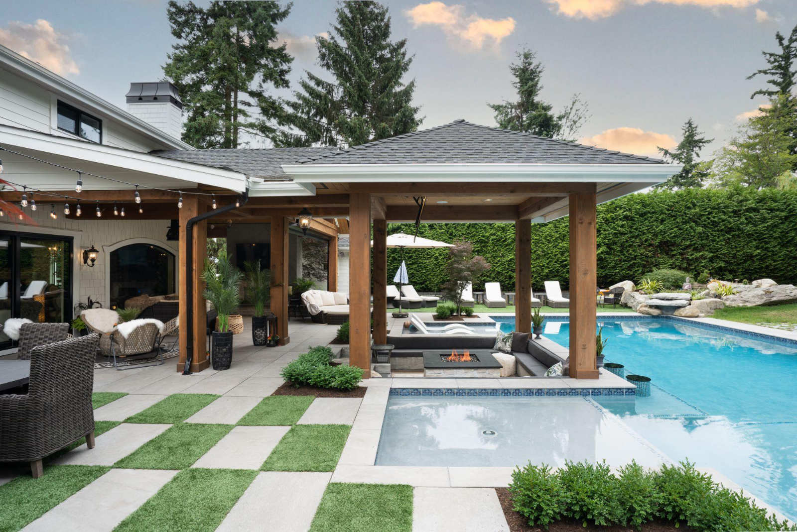 High-end outdoor living space of a Metro Vancouver forever home, featuring a luxury swimming pool, a covered patio with a sunken lounge and fire pit, a stone-paved deck, and a lush green privacy hedge.