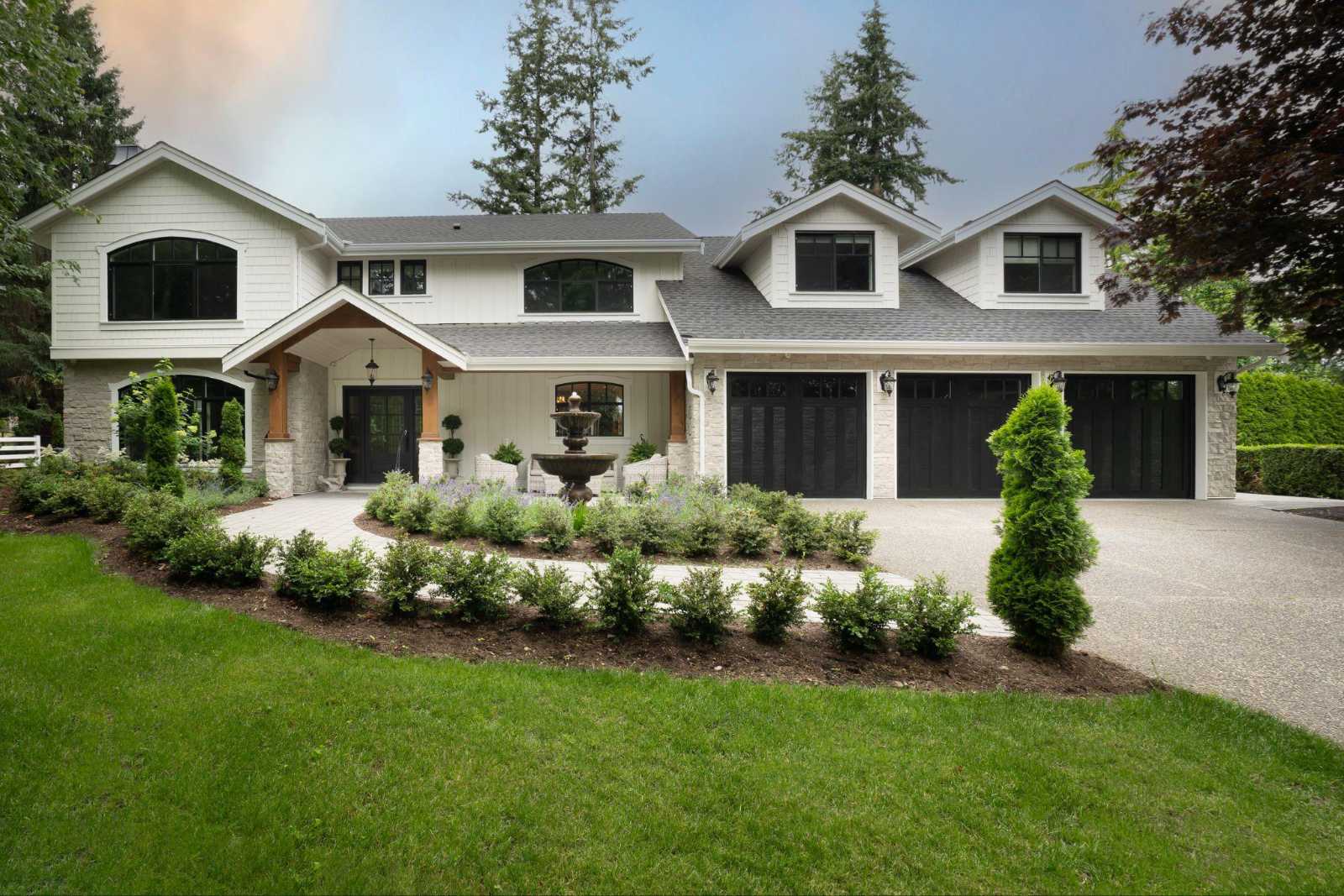 custom-home-design-abbotsford-forever-home Exterior front view of a luxury modern farmhouse custom home in Abbotsford, featuring a long paved driveway, immaculate landscaping with a central water fountain, white siding, stone accents, and a three-car garage.