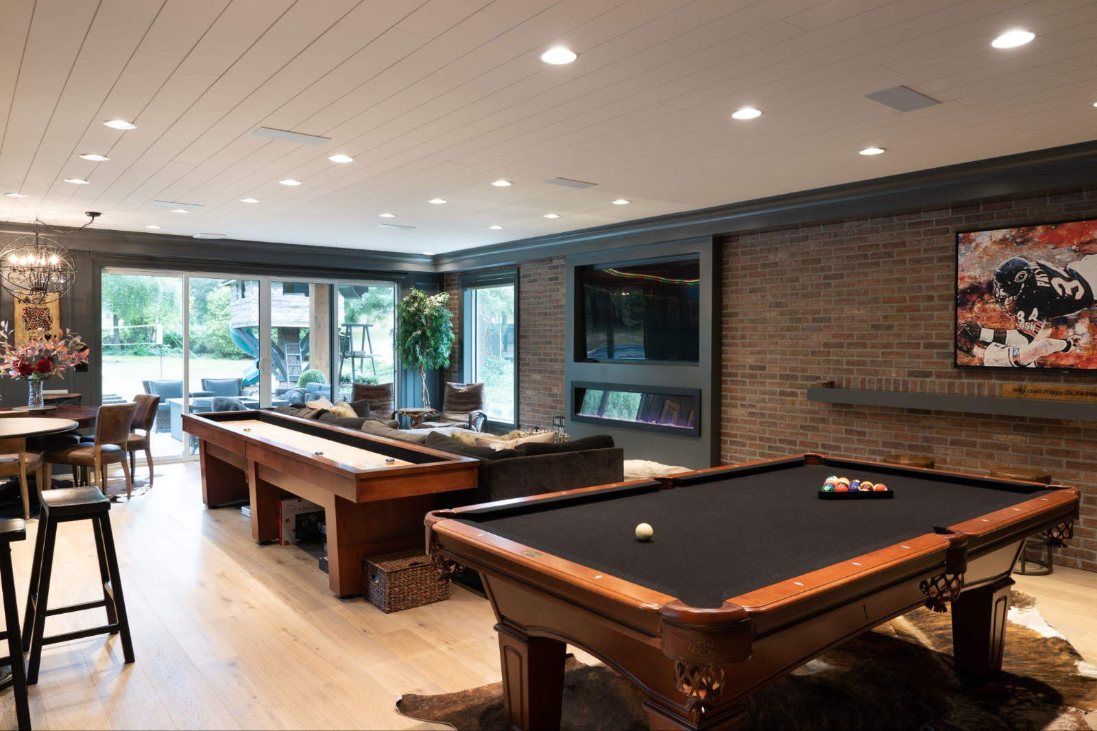 Spacious basement entertainment layout from a high-end home renovation, showcasing an open-concept game room with a pool table, shuffleboard, a seating area with a brick accent wall, and sliding glass doors to the backyard.