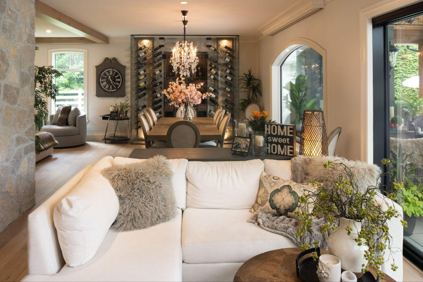 Warm and inviting open-concept living and dining area in a Fraser Valley custom home, featuring a white sectional sofa, a rustic dining table beneath a crystal chandelier, a glass wine display wall, and natural stone pillars.