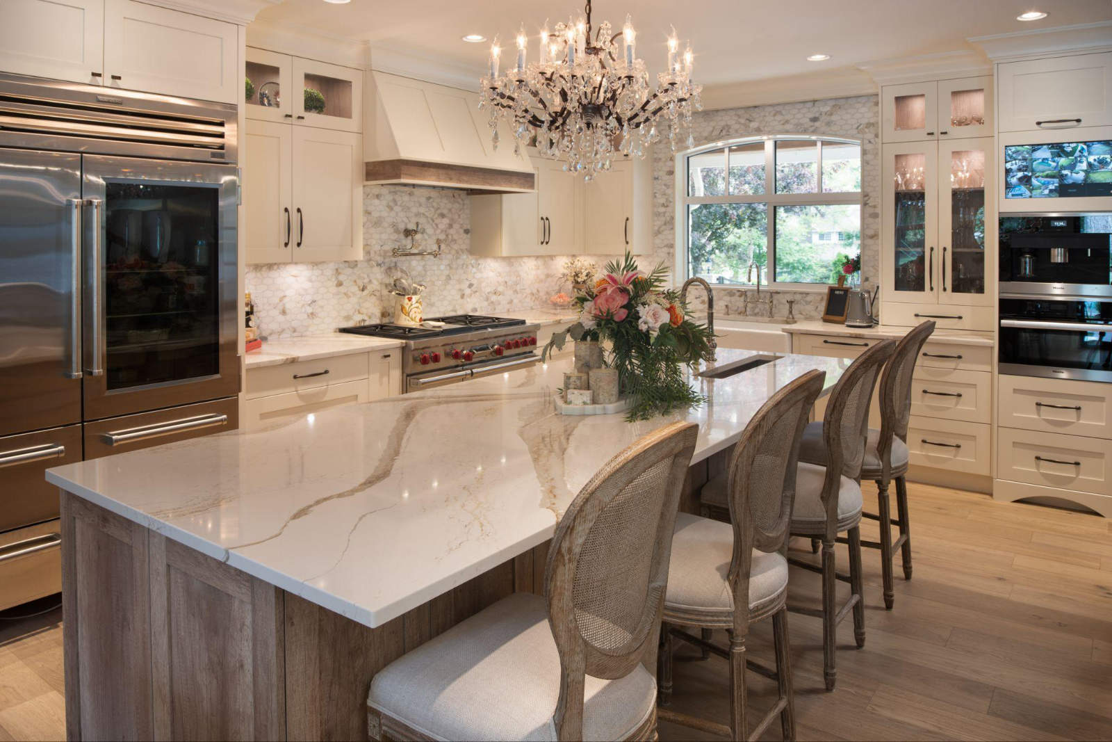 Timeless interior design 2026 kitchen featuring a large marble island with gold veining, elegant wooden bar stools, cream-colored custom cabinetry, high-end stainless steel appliances, and a crystal chandelier.