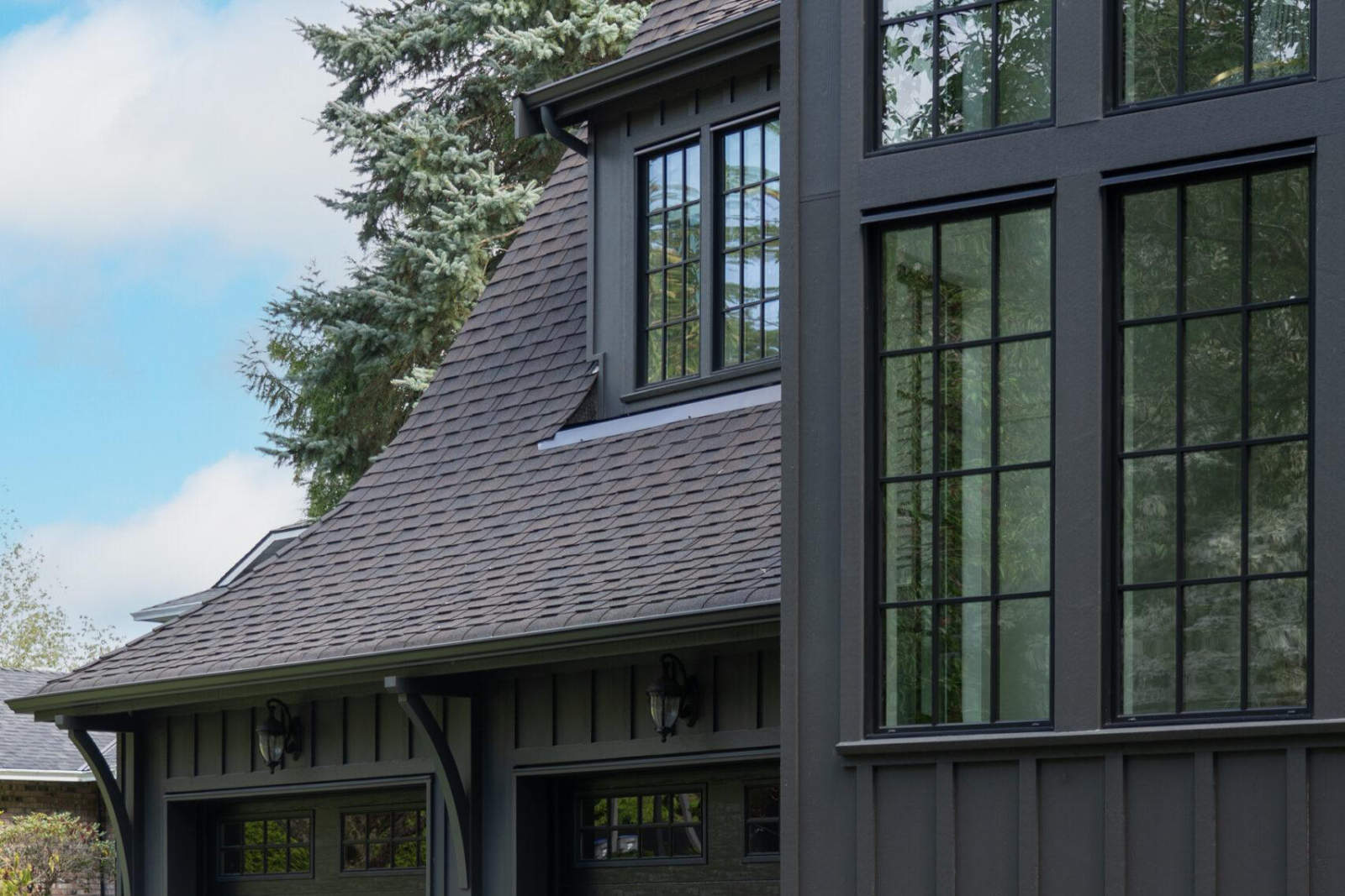 Architectural detail of a dark gray luxury home exterior and shingled roofline near mature evergreen trees.