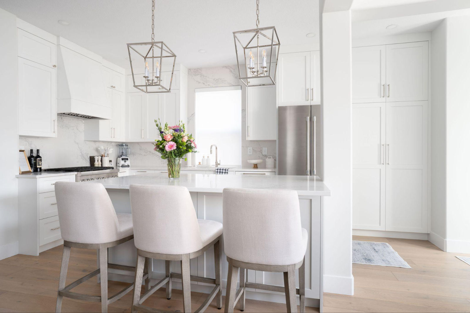 Spacious white custom kitchen featuring a large island, upholstered bar stools, cage pendant lights, and full-height cabinetry.