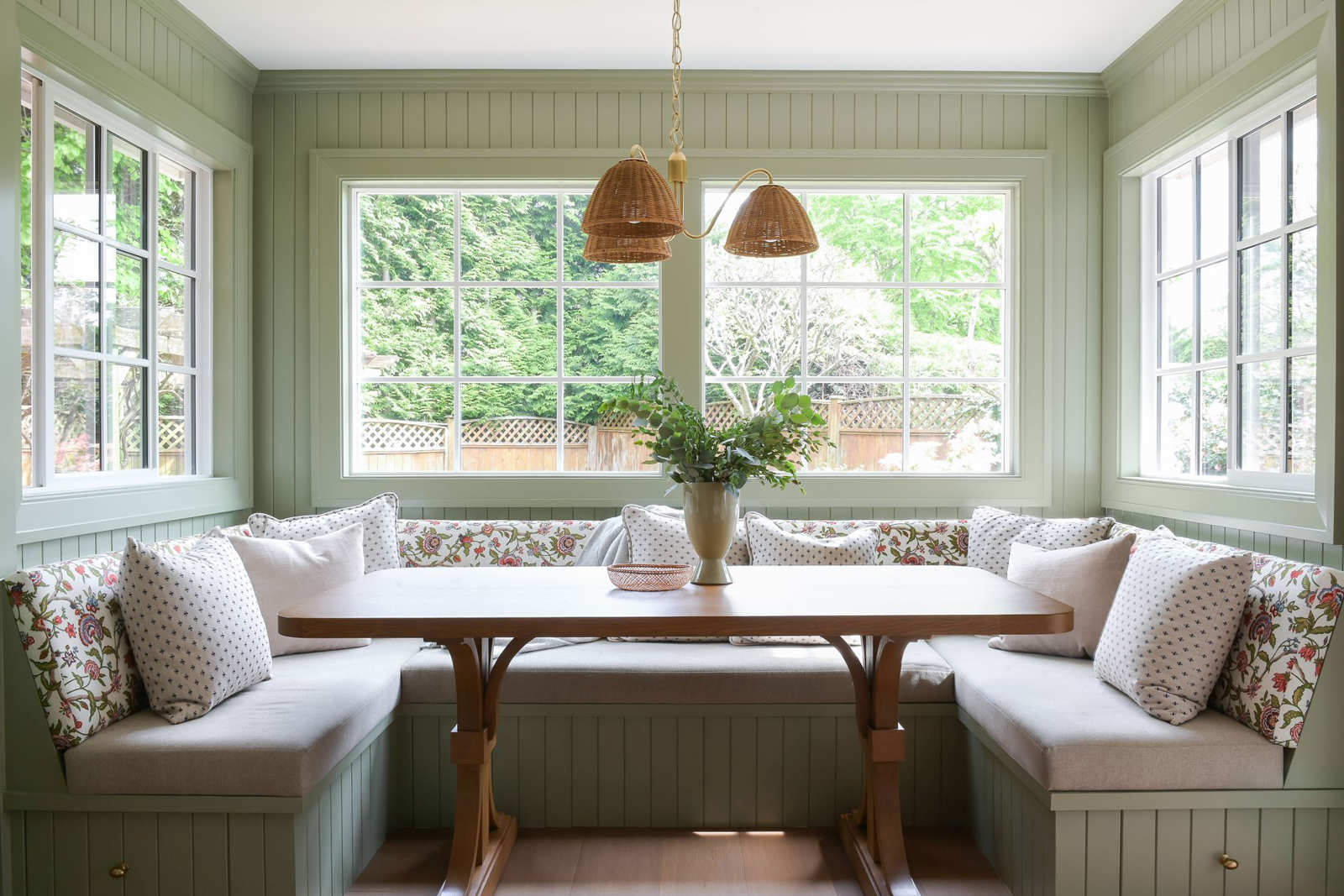 A cozy built-in banquette dining nook with sage green paneling, floral pillows, and large windows looking out onto a backyard garden.