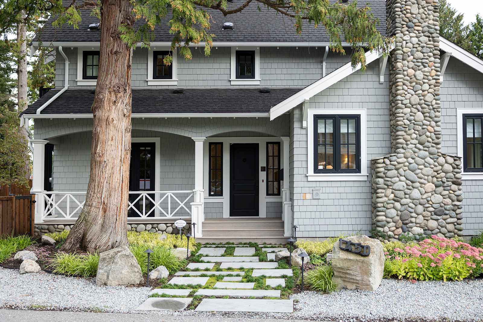 Front entrance of a Craftsman-style home showing grey shingle siding, a stone chimney, a stone walkway, and front porch.