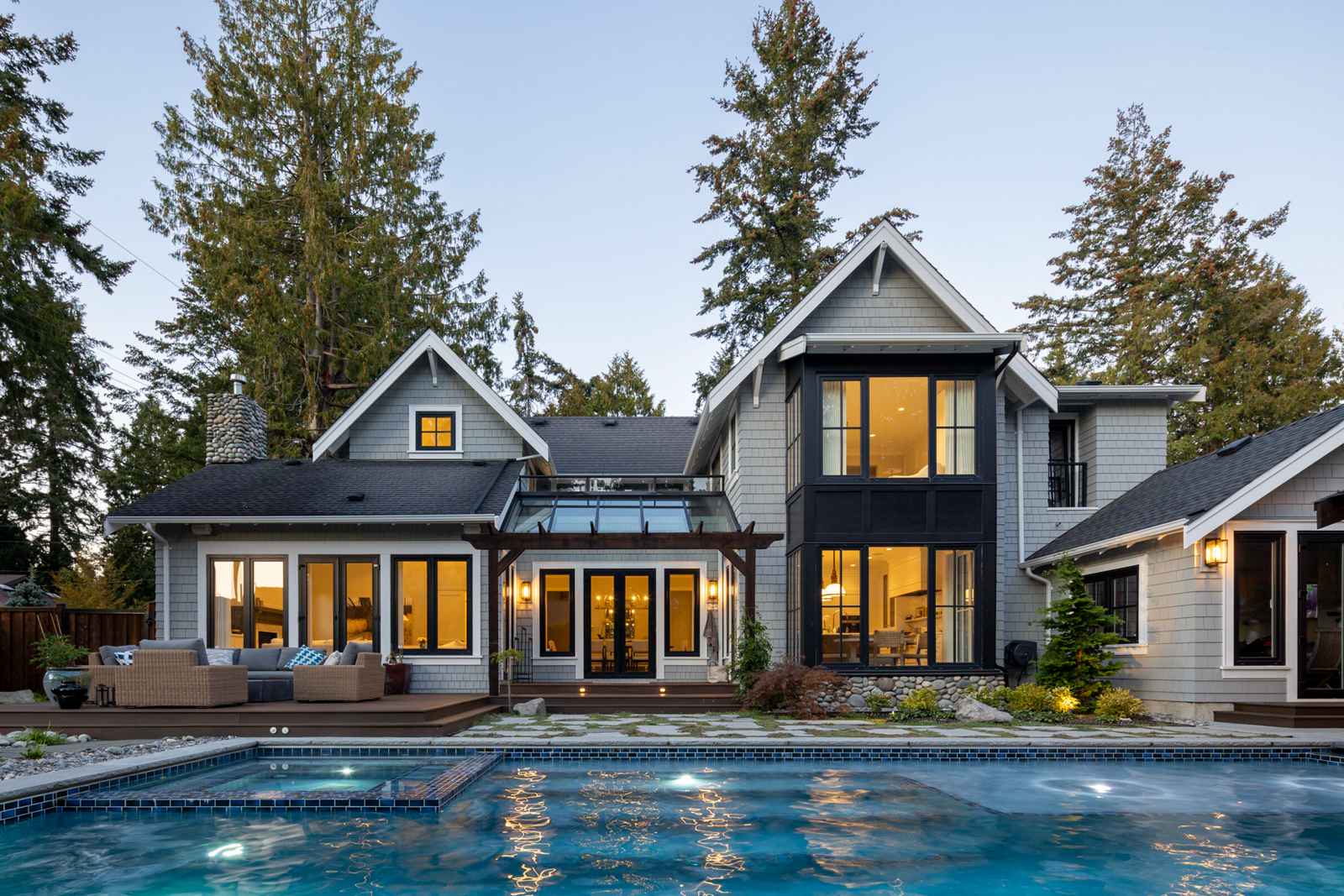 Rear view of a luxury custom forever home featuring a back patio, illuminated pool, and large glass windows surrounded by mature trees.