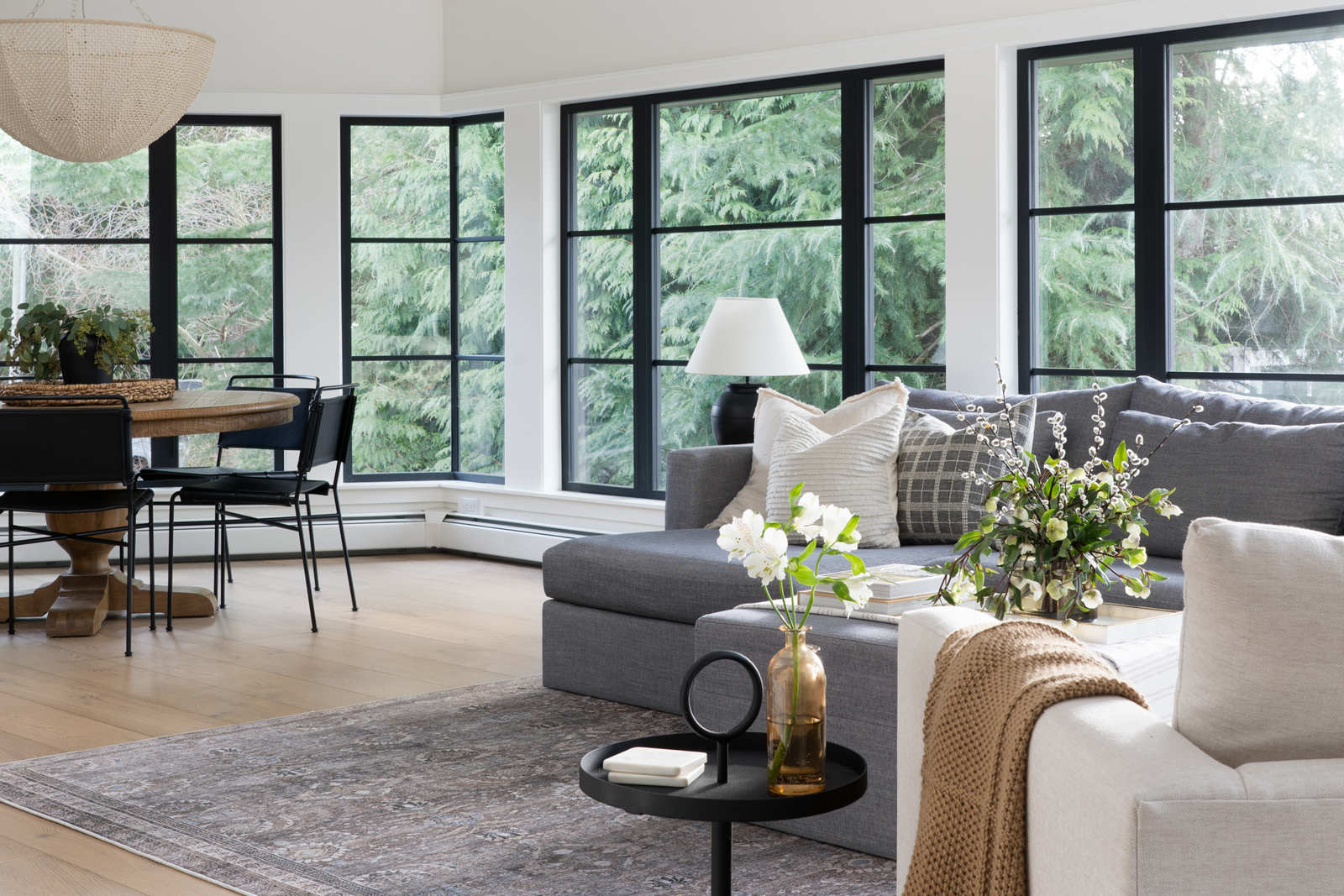 A bright, open-concept living space with floor-to-ceiling black window frames, a gray sectional sofa, and a cozy round dining area.
