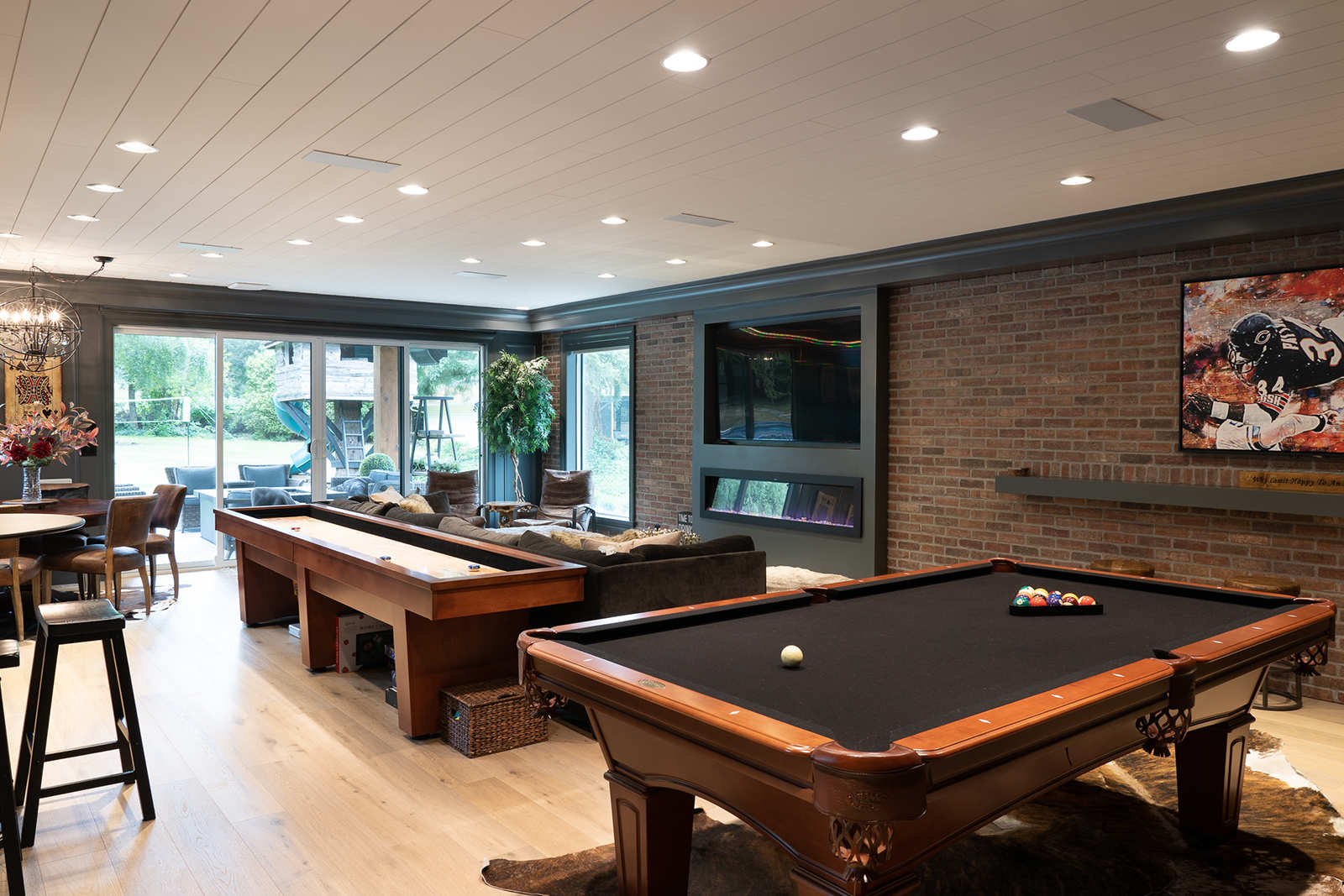 Spacious basement game room designed for family gatherings, featuring a pool table, shuffleboard table, lounge area, and brick wall accents.