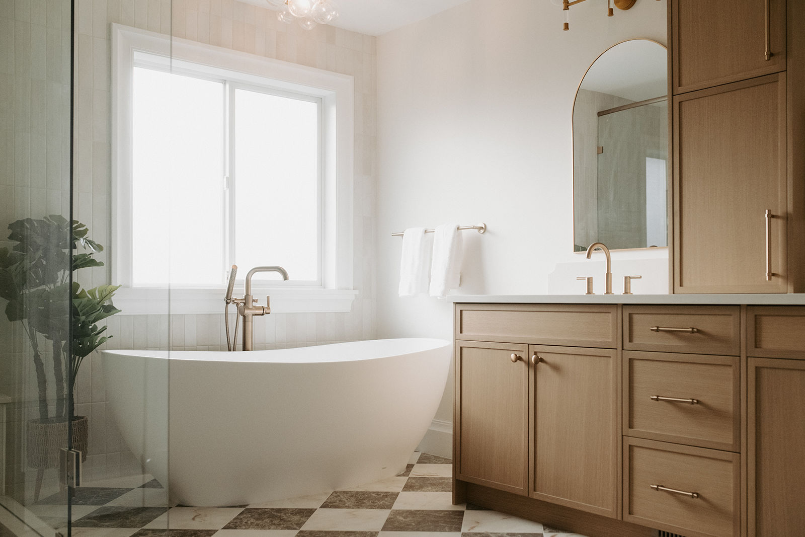 White freestanding soaking tub positioned next to a window in a bright bathroom with light wood cabinets and checkered flooring.