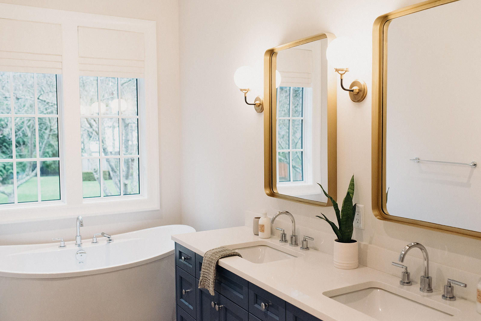 Luxury primary bathroom featuring a dark blue double vanity with brass mirrors, brass sconces, and a white soaking tub by the window.